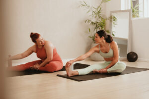 Two females exercising together in fitness studio. Women in sports clothing doing leg stretching workout at gym.